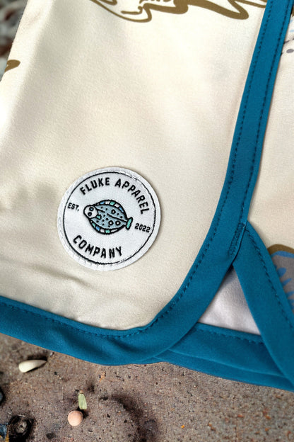Beige fabric with blue trim and Fluke Apparel Company logo on a sandy background